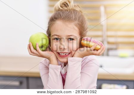 Cute little girl choosing between apple and donut in the kitchen