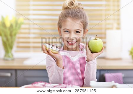 Cute little girl choosing between apple and donut in the kitchen
