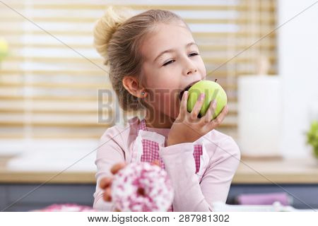 Cute little girl choosing between apple and donut in the kitchen