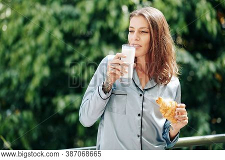 Beautiful Young Woman In Pajamas Having Glass Of Milk Or Yogurt And Croissant For Breakfats When Sta