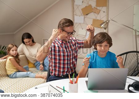 Happy Dad And Son Sitting At The Table At Home, Looking At Laptop And Celebrating Success While Moth