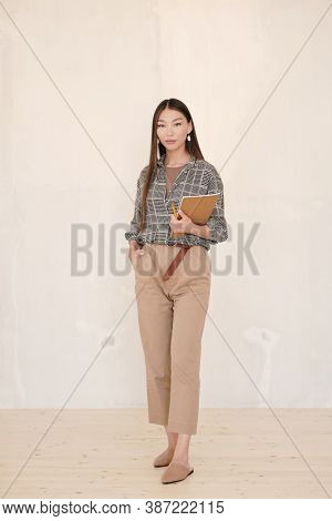 Young businesswoman with dark long hair holding notebook and pencil while standing in front of c amera against white wall in studio