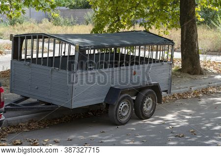 Two-axle Car Trailer On The Roadside. Empty Metal Trailer With A Roof In The Shade Of Trees On A Cit