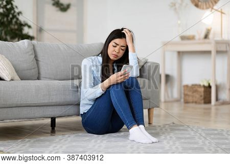 Waiting For His Call. Unhappy Korean Woman Looking At Smartphone Screen At Home While Sitting On Flo