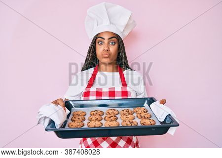 Young african american woman with braids wearing baker uniform holding homemade cookies puffing cheeks with funny face. mouth inflated with air, catching air. 