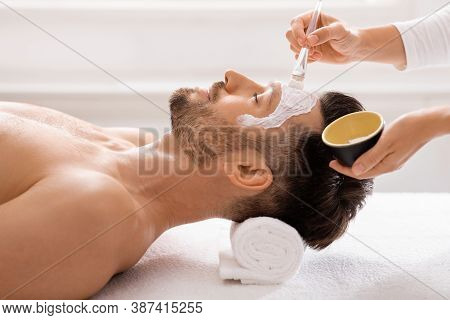 Side View Of Bearded Man Getting Face Treatment At Modern Spa Salon. Female Attendant Applying Nouri