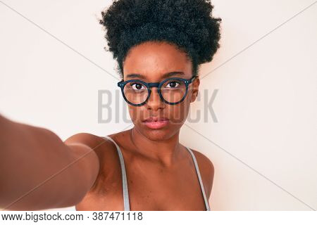Young african american woman wearing casual clothes and glasses making selfie by the camera thinking attitude and sober expression looking self confident 