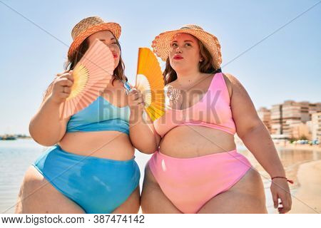 Two plus size overweight sisters twins women using handfan at beach on hoy day of summer