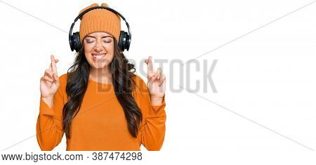 Beautiful brunette young woman listening to music using headphones gesturing finger crossed smiling with hope and eyes closed. luck and superstitious concept. 