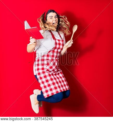 Young beautiful blonde cook woman wearing apron smiling happy. Jumping with smile on face holding recipe book and spoon over isolated red background
