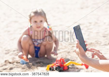 Hands Of A Woman Photographing A Small Three-year-old Girl On Her Phone Playing With A Machine On Th