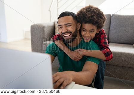 Mixed race father with son enjoying family time together at home, social distancing and self isolation in quarantine lockdown, boy is embracing man from the back, using laptop and smiling