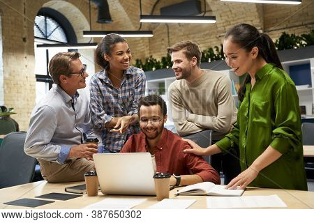 Group Of Young Happy Business People Looking At Laptop Screen, Communocating And Discussing Work Whi