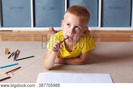 A Cute Blond Boy In A Yellow T Shirt Is Lying On The Floor With A Sheet Of Paper And Pencils And Loo