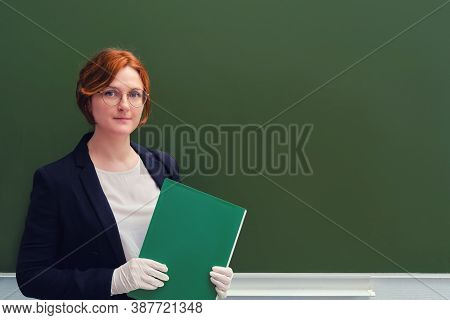 Woman Teacher Holds A Green Folder While Standing Near The School Blackboard, Copy Space