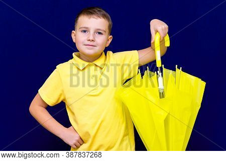 Attractive Surprised Caucasian Blond Schoolboy Holds Unexpanded Umbrella On Blue Studio Background.b
