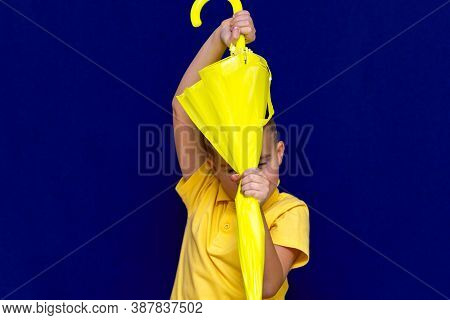 Attractive Surprised Caucasian Blond Schoolboy Holds Unexpanded Umbrella On Blue Studio Background.b