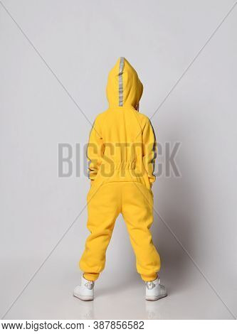 Full-growth Portrait Kid Girl In Stylish Yellow Jumpsuit And Sneakers Stands Back To Camera With Her