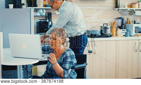 Disabled Old Woman In Wheelchair Working On Laptop In Kitchen. Paralysied Handicapped Old Elderly Pe