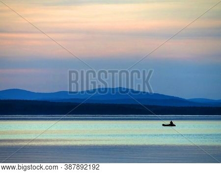 Calm Blue Sunset Sky Over The Lake, Silhouette Of A Lone Boat With A Fisherman, Calm Surface Of Wate
