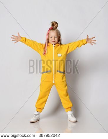 Blonde Kid Baby Girl With Colorful Braids Wearing Stylish Yellow Jumpsuit And Sneakers Stands With H