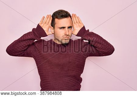 Young handsome man with blue eyes wearing casual sweater standing over pink background Trying to hear both hands on ear gesture, curious for gossip. Hearing problem, deaf