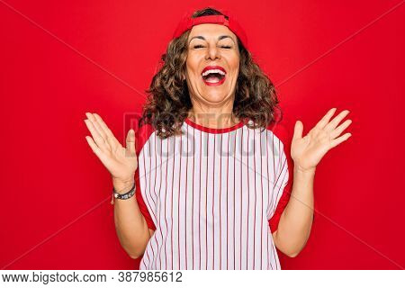 Middle age senior woman wearing baseball equiment over red isolated background celebrating mad and crazy for success with arms raised and closed eyes screaming excited. Winner concept