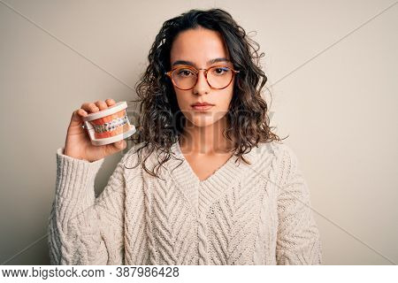 Young beautiful woman with curly hair holding plastic teeth with dental baces with a confident expression on smart face thinking serious