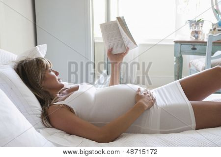 Genç hamile kadın yatakta yatarken kitap okumak yan görünüm
