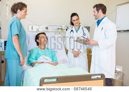 Young doctors discussing notes while happy patient and nurse looking at them in hospital room