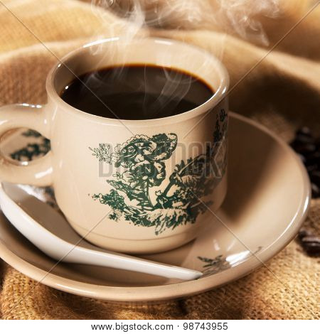 Steaming traditional oriental Chinese kopitiam style dark coffee in vintage mug and saucer with coffee beans. Fractal on the cup is generic print. Soft focus with dramatic light on wooden background.