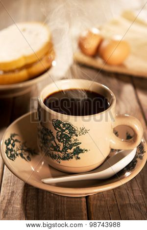 Steaming traditional oriental Chinese kopitiam style dark coffee in vintage mug and saucer with coffee beans. Fractal on the cup is generic print. Soft focus with dramatic light on wooden background.