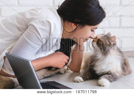Young woman and her lovely cat rubbing noses each other. Fluffy family pet lays on working table with laptop nearby