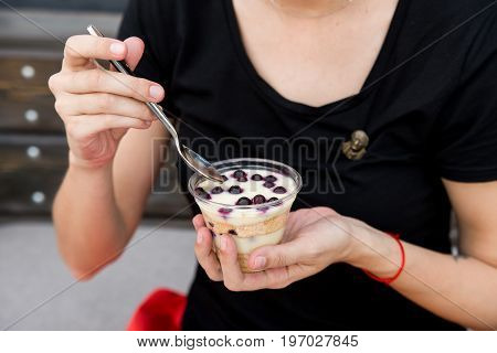 Close up female hands holding spoon and plastic glass with delicious dessert with blueberries and custard