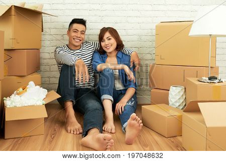 Full length portrait of cheerful Asian couple posing for photography while sitting on floor of living room surrounded with moving boxes