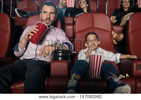 Handsome Hispanic young man and his son sitting in the movie theather and enjoying a film together
