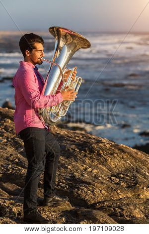 Tuba - instrument. Young man play the trumpet on rocky sea coast during surf.