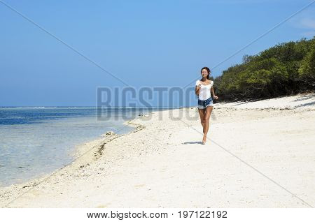 Active sporty woman run along ocean surf by water pool. Sunset sand beach background with blue sky and sun. Woman fitness, jogging workout and sport activity on summer holiday.