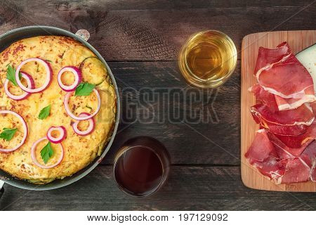 An overhead photo of a Spanish tortilla with potatoes and zucchinis in a tortillera, with jamon, cheese, glasses of white and red wine, with a place for text