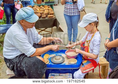 Zaporizhia/Ukraine- May 28, 2017: Charity Family festival: Outdoors pottery workshop - ceramist master explaining  girl how to hold fingers, making clay jug . Art and craft activity.