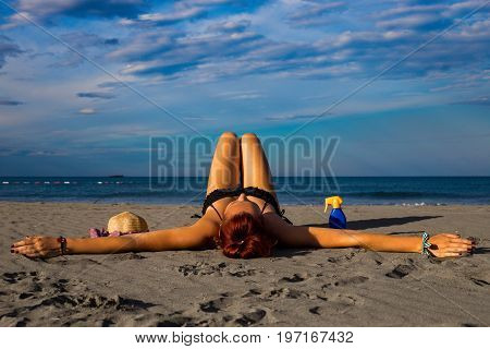 Young good looking tanned redhead laying on a sandy beach with arms resting and leg resting and happily posing with arms widespread with sun cream and hat on the side and heavenly blue sky and sea in the background during summer