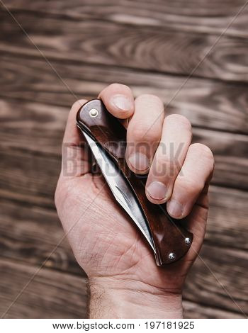 Male hand holding stainless jackknife on a wooden background.