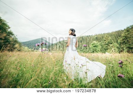 Beautiful young woman wearing elegant white dress standing on a feild and looking to the mountains