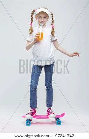 Full Length Portrait of Happy Caucasian Blond Girl in Visor Ballancing on Pennyboard With Cup of Juice.Against White. Vertical Image Composition