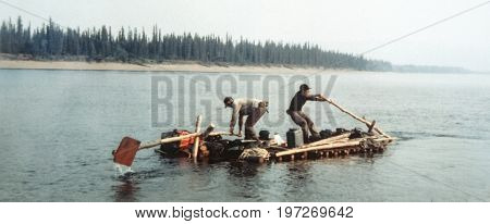 Two men rowing down the riveron self-made log raft. Vintage expedition like in old gold rush times. Alaska. Blurry and grainy old-style photography.
