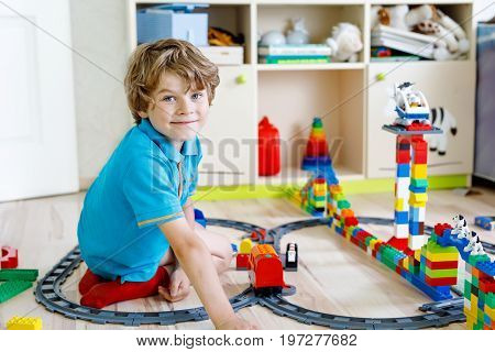 Adorable little blond kid boy playing with colorful plastic blocks and creating train station. Child having fun with building toys at home.