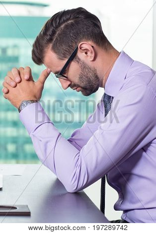 Closeup of thoughtful young business man leaning head on fingers, relaxing and sitting at table with big window and building view outside in background. Side view.