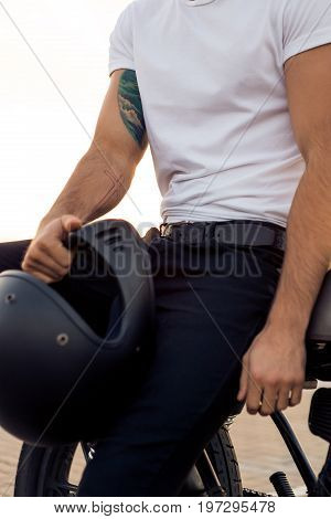Close up of a handsome rider male in white blank t-shirt with helmet near classic style cafe racer motorcycle at sunset. Bike custom made in vintage garage. Brutal urban lifestyle. Outdoor portrait.