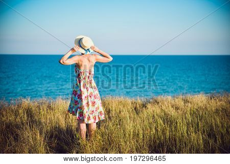 Summer Portrait Of Young Pretty Woman In A Straw Hat And Dress Standing Backwards In The Grass And L