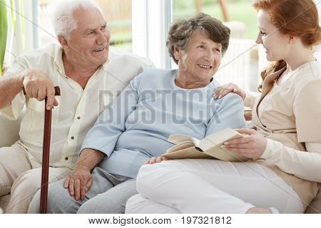 Two elder people are looking at smiling nurse who reads book in common room